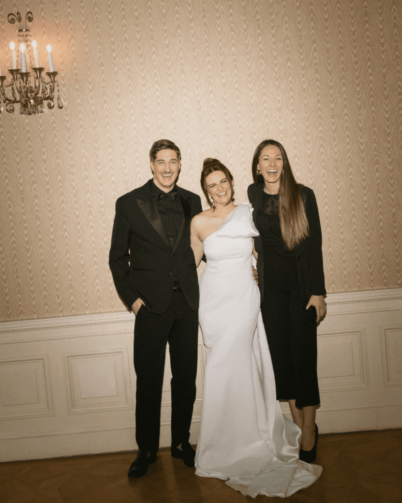 Brautpaar posiert gemeinsam mit Hochzeitsplanerin in elegantem Innenraum und lacht in die Kamera