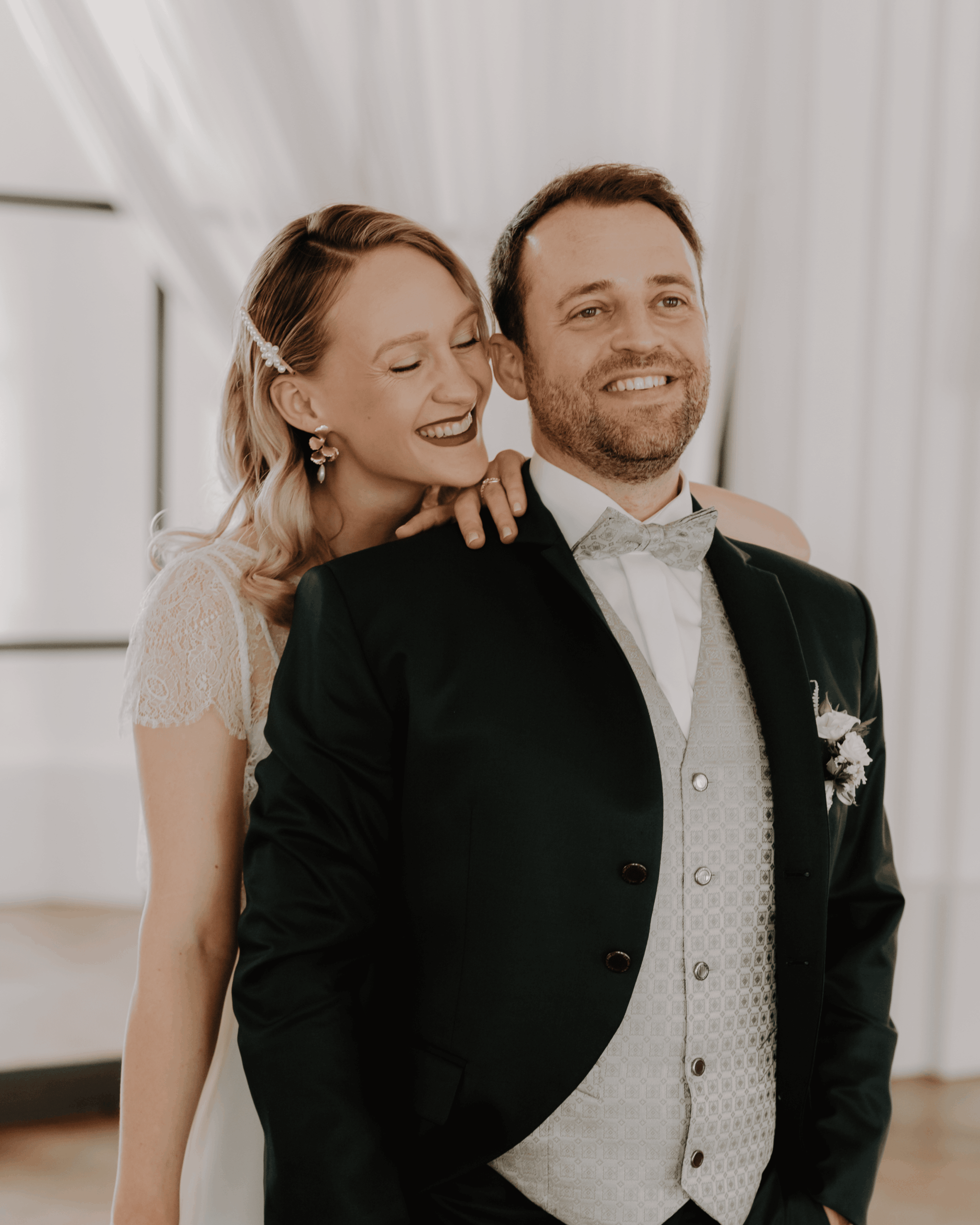 Braut lehnt sich lächelnd an den Bräutigam, emotionales Hochzeits-Portrait in hellem Innenraum